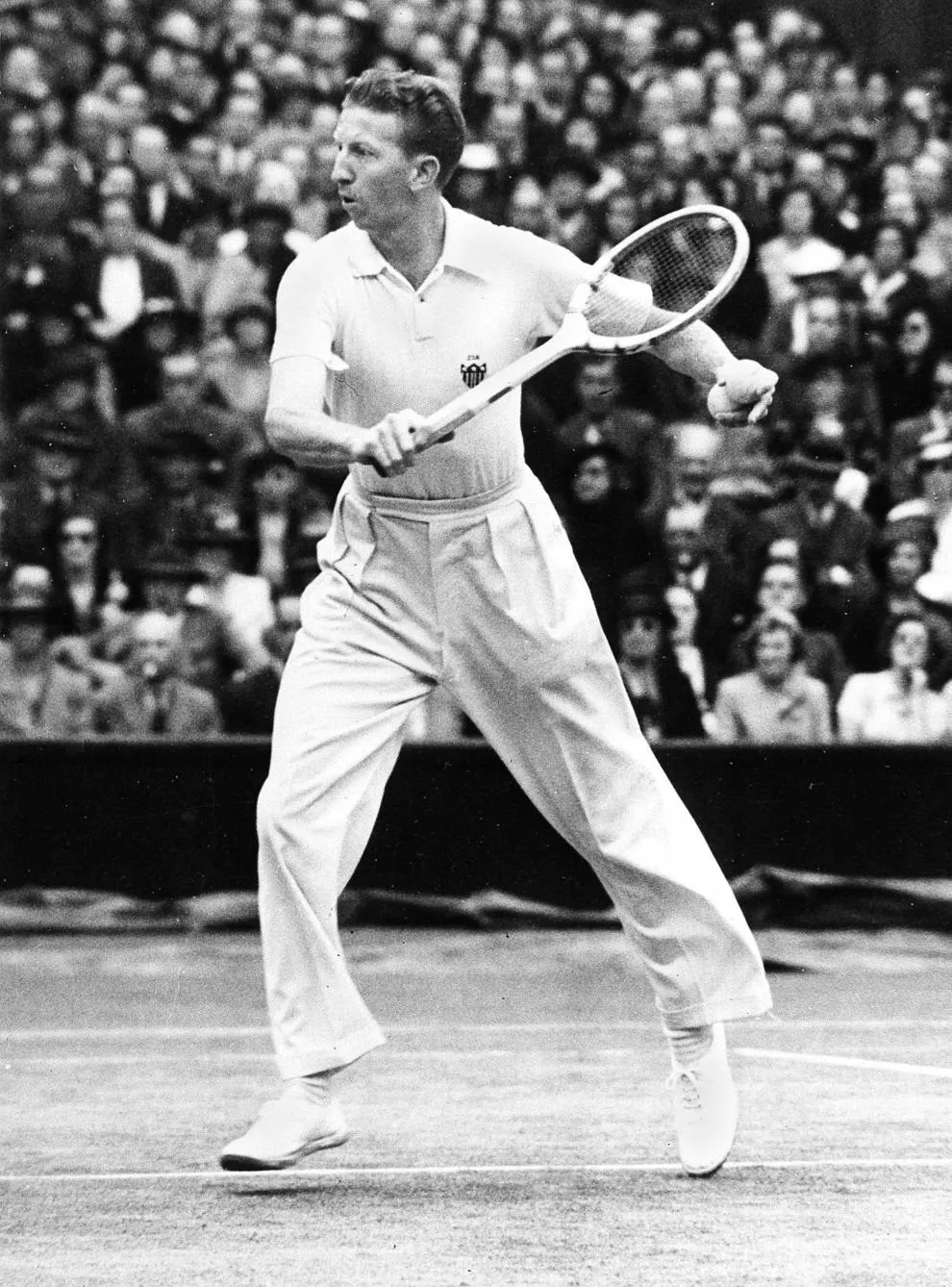 Black-and-white photo of Don Budge in tennis whites mid-stroke in front of a packed grandstand